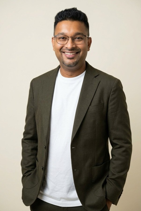 Dinesh Dananjaya smiling, wearing glasses, a white t-shirt and an olive blazer